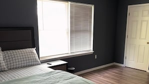 A bedroom with a bed, a nightstand, a window with blinds, a door, and wooden flooring. - NWK 457  ROOM 211-D (Newark)
