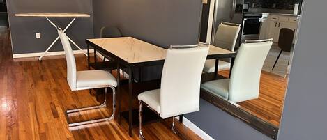 Un espace repas moderne avec une table en bois et des chaises blanches, une petite table pliante et une cuisine en arrière-plan.