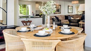 A dining area with a round wooden table surrounded by wicker chairs, a vase with flowers, and a view of a kitchen and living area in the background.