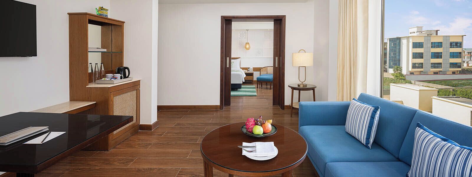 Une chambre d’hôtel avec un canapé, une table basse sur laquelle se trouvent des fruits, un téléviseur, un meuble en bois et une vue sur les immeubles.