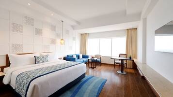 A modern hotel room with a large bed, a blue sofa, a small table, and a window with curtains.