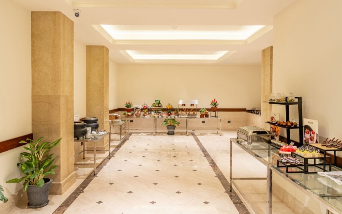 Een hotel lobby met een marmeren vloer, een receptiebalie, een buffetgedeelte met diverse etenswaren en potplanten.