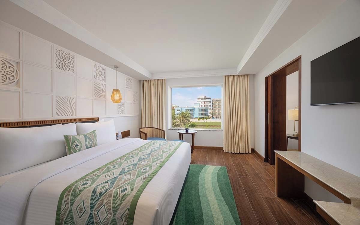 A hotel room with a bed, a desk, a chair, a TV, a window with curtains, and a view of buildings.