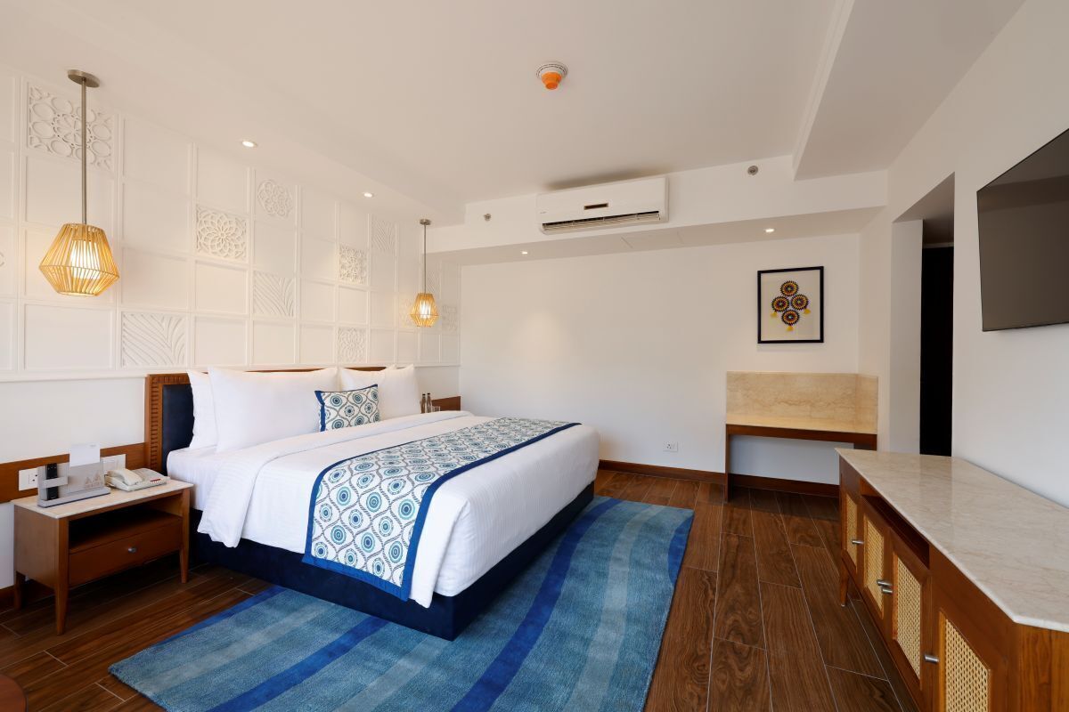 A hotel room with a large bed, a bedside table, a desk, a chair, a TV, and a wall-mounted air conditioning unit.