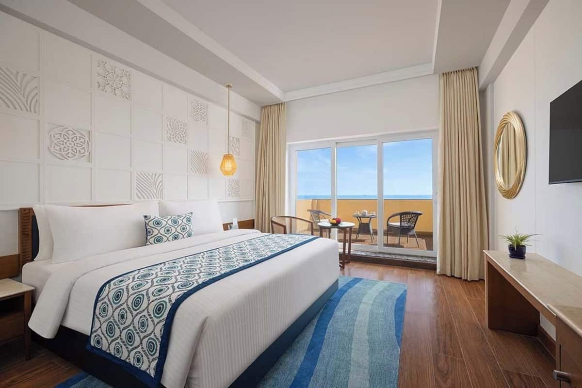A hotel room with a large bed, a desk, a chair, a TV, and a balcony with a view.