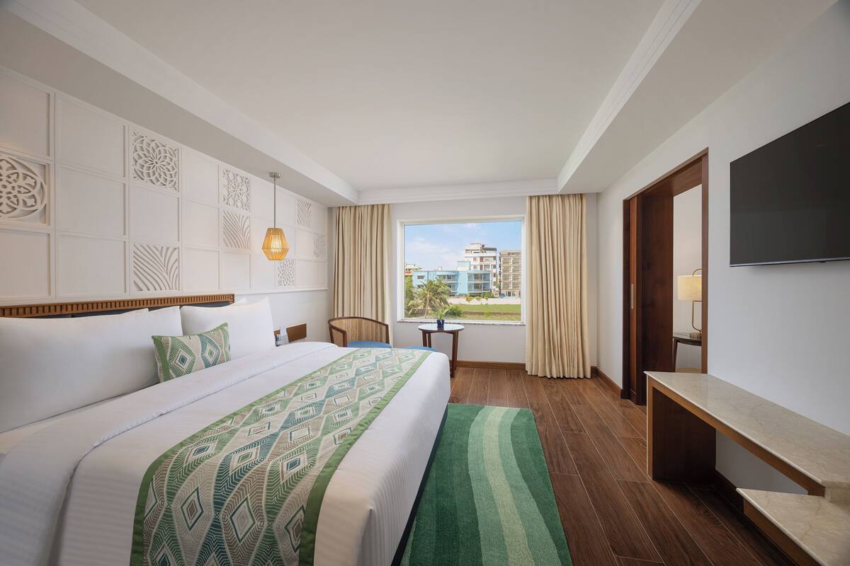 A hotel room with a large bed, a desk, a TV, and a window with a view of buildings.