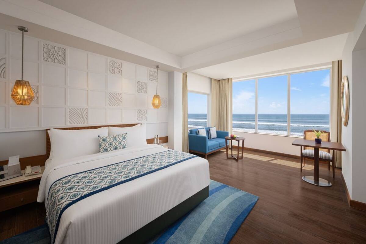 A hotel room with a large bed, a view of the ocean, a seating area with a table and chairs, and a decorative wall panel.