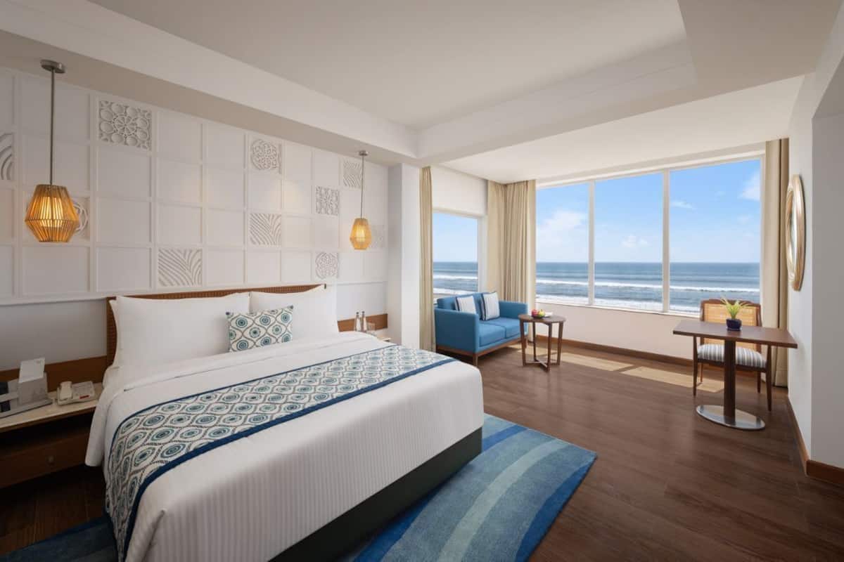 A hotel room with a large bed, a view of the ocean, a seating area with a table and chairs, and a decorative wall panel.