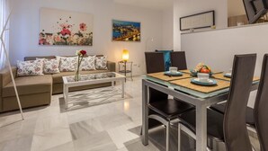 Un salon moderne comprenant un canapé, une table basse, une table à manger et des chaises. Deux tableaux ornent le mur, ainsi qu’une lampe et un téléviseur fixé au mur.