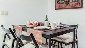 Ein Esstisch mit rot-weiß kariertem Tischläufer, Tellern, Gläsern und einer Schüssel mit Tomaten. Stühle stehen um den Tisch, und ein gerahmtes Poster ist an der Wand zu sehen.