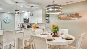 A modern kitchen with white cabinetry, a dining area with white chairs, and a wall-mounted surfboard sign. - Pool & Beach Trolley Access: PCB Resort Condo! (Panama City Beach)
