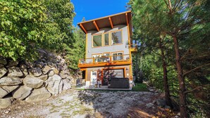 Une cabane à deux étages avec une entrée en pierre, une terrasse et une grande fenêtre donnant sur une zone boisée.