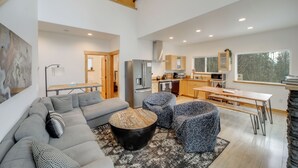 Un salon moderne avec un canapé d’angle, une table basse ronde et des fauteuils à motifs. On aperçoit à l’arrière-plan une cuisine avec un coin repas et de grandes fenêtres.