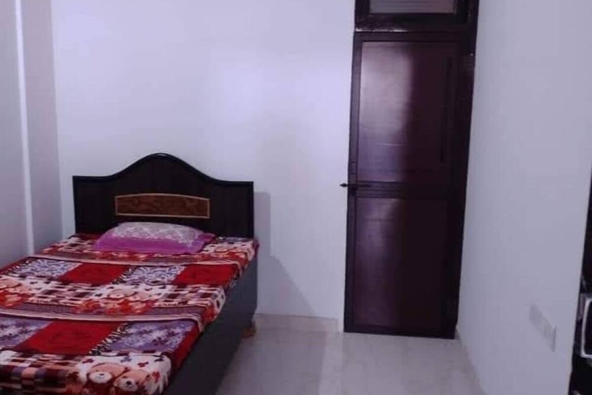 Ett litet, smalt rum med en enkel säng, en mörk dörr och en takfläkt.