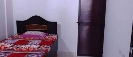 A small, narrow room with a single bed, a dark door, and a ceiling fan.