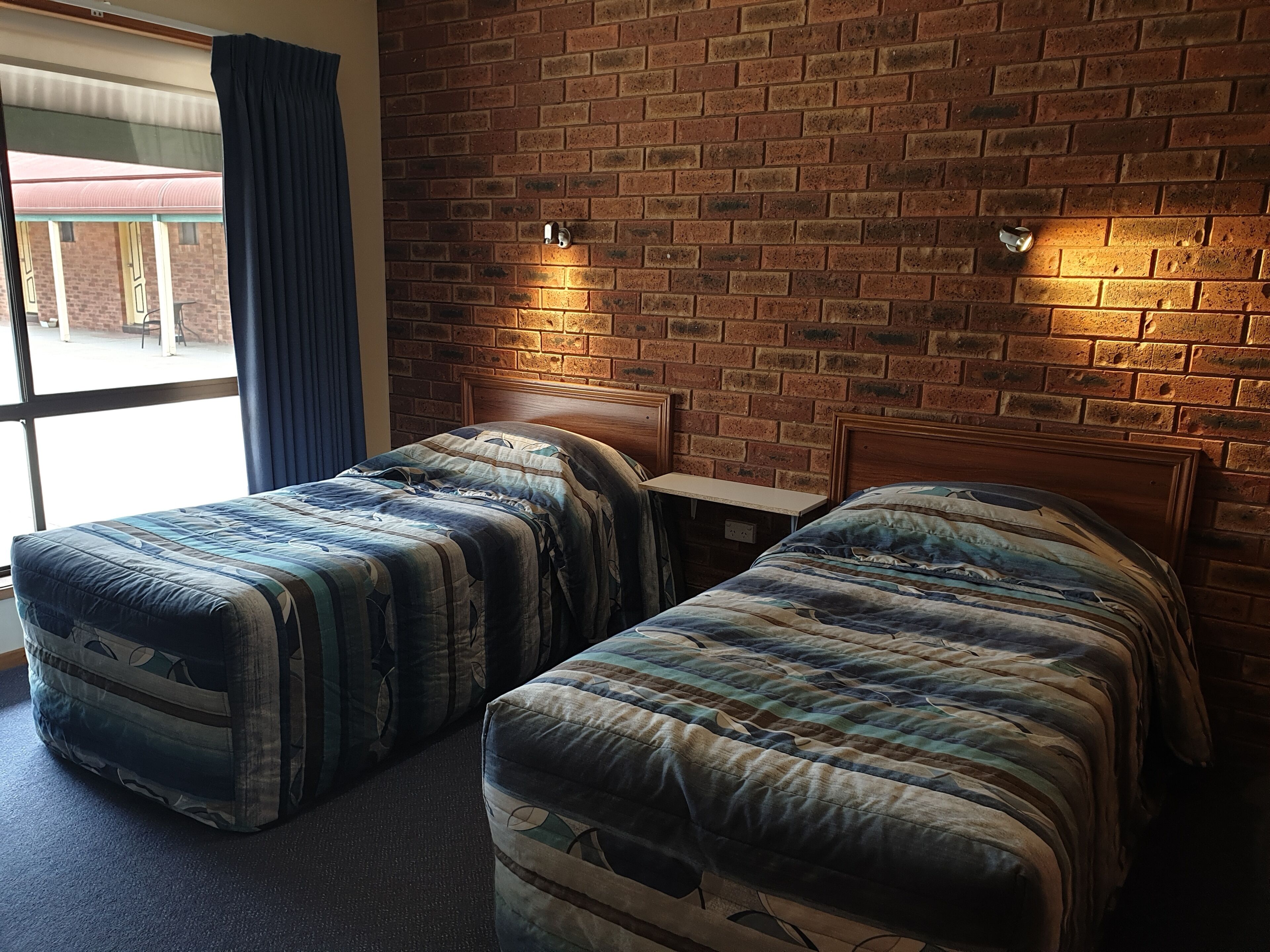 Ein Hotelzimmer mit zwei Einzelbetten, einem Holzkopfende, einer Ziegelwand und einem Fenster mit Vorhängen.