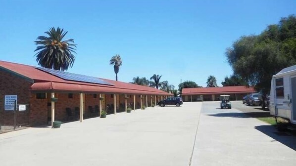 A row of motel-style buildings with red roofs and solar panels, a parking area, and a trailer park to the right.