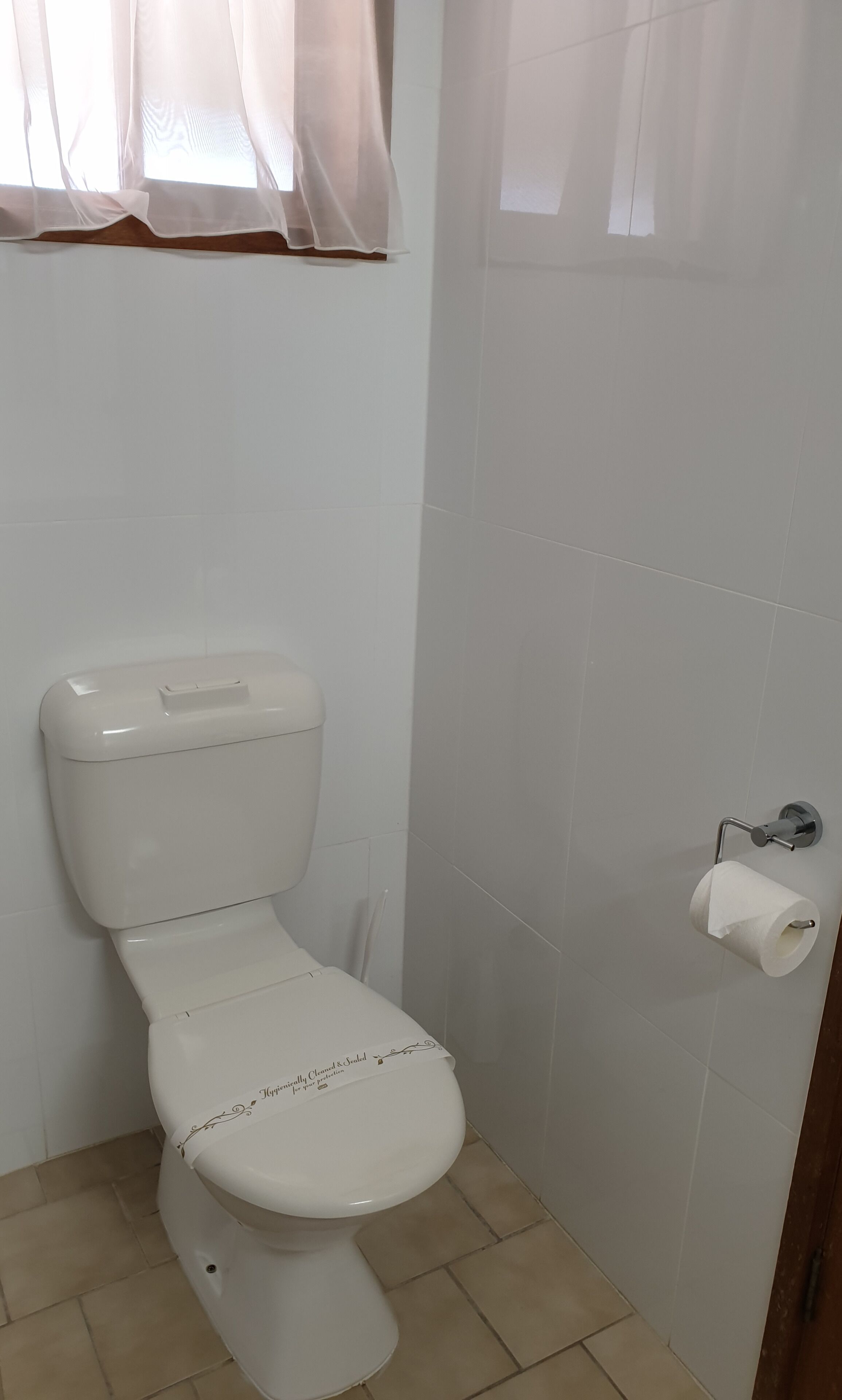 A white toilet with a lid, a toilet paper holder, and a window with sheer curtains.