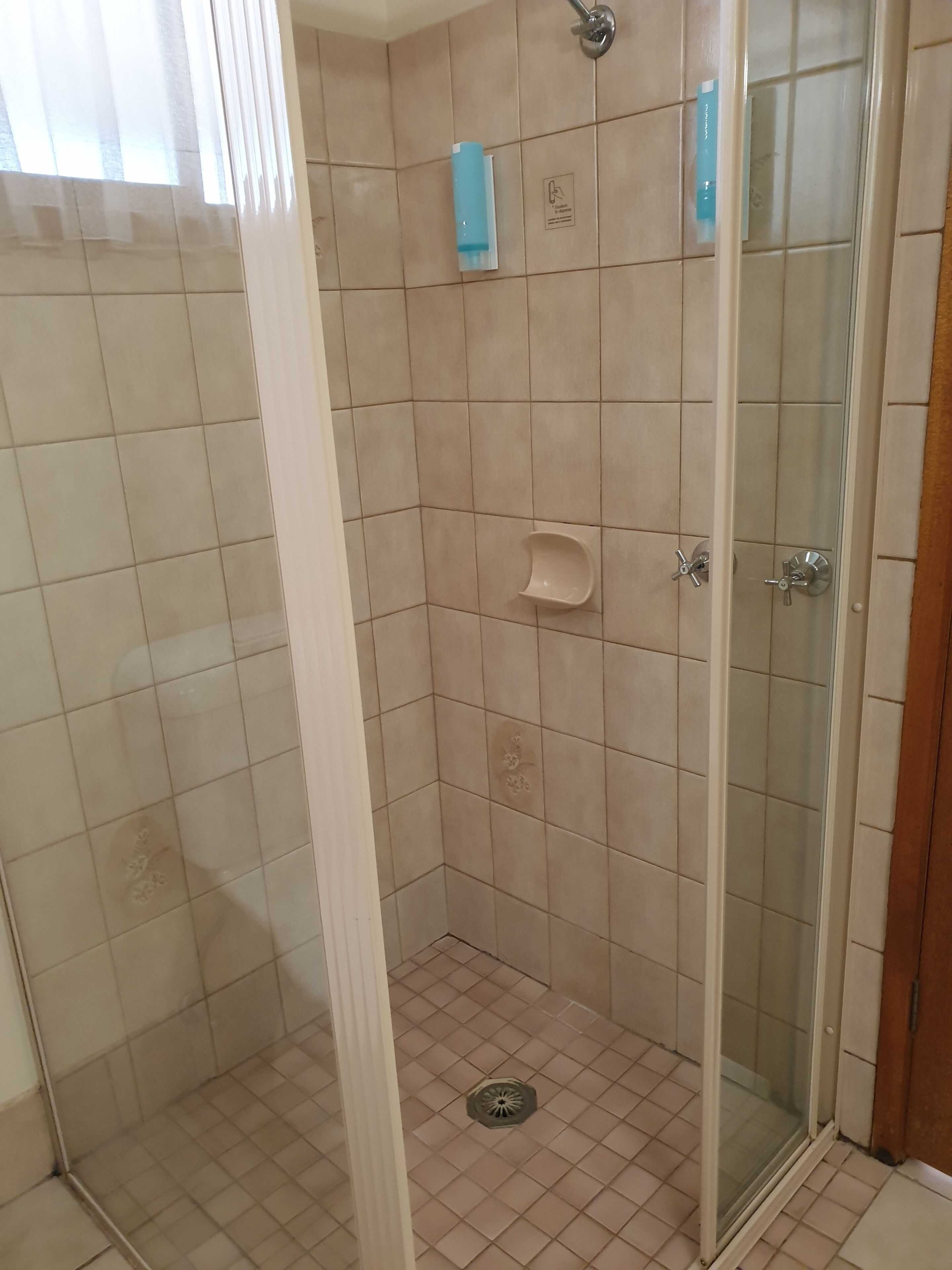 Un petit espace de douche carrelé, doté d’une porte en verre, d’un distributeur de savon liquide et d’un pommeau de douche.