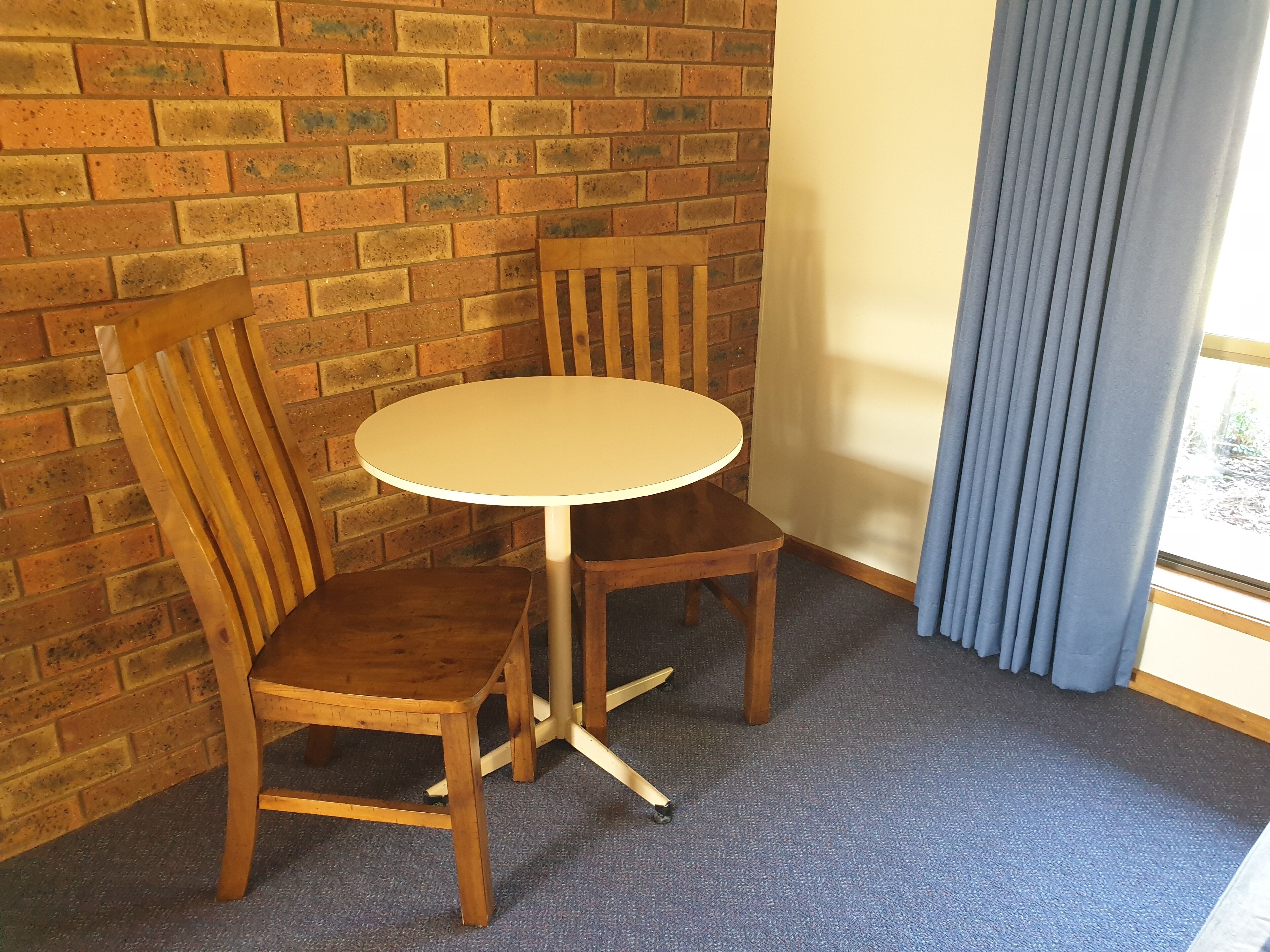 Une petite pièce avec une table ronde et deux chaises en bois, un mur en briques et une fenêtre avec des rideaux.