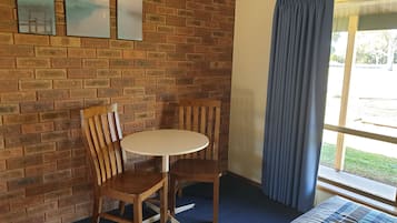 A room with a brick wall, two wooden chairs, a small round table, a bed with a blue and white checkered bedspread, and a window with blue curtains.