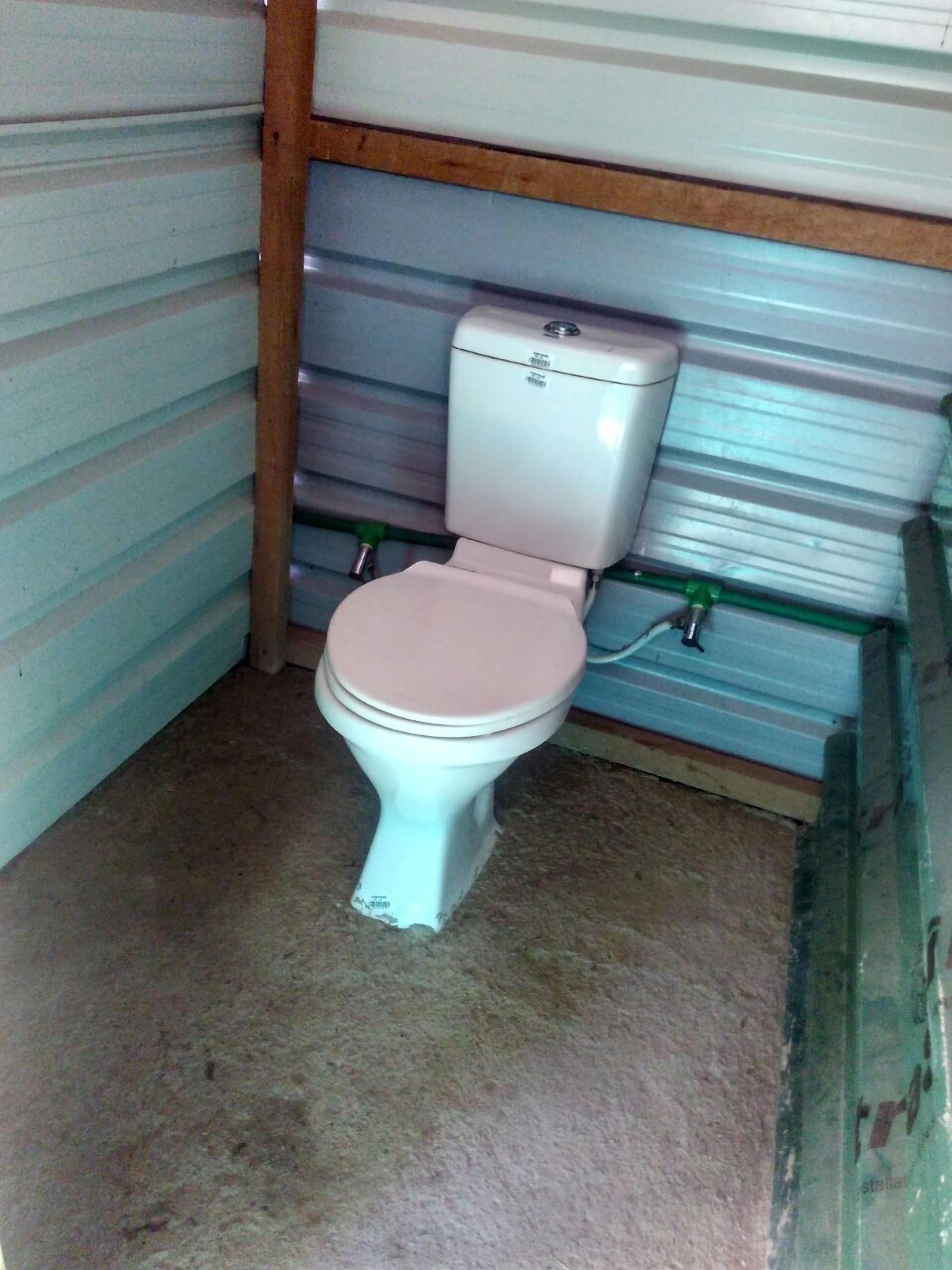 Ein kleines, abgeschlossenes Badezimmer mit Toilette, einer grünen Leitung und einem Betonboden.