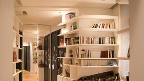 Een smalle gang met witte wandplanken vol boeken en decoratieve voorwerpen, die leidt naar een donkere deur.