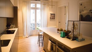 Een moderne keuken met een centraal eiland, een eethoek met een witte tafel en stoelen, en een uitzicht door een groot raam met gordijnen.