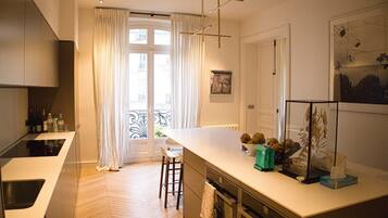 Eine moderne Küche mit einer zentralen Kochinsel, einem Essbereich mit weißem Tisch und Stühlen und Blick durch ein großes Fenster mit Vorhängen.