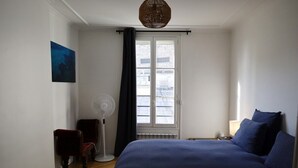 A bedroom with a bed, a chair, a standing fan, and a window with curtains. - Beautiful apartment 4 ppl. / Pigalle - Montmartre (Paris)
