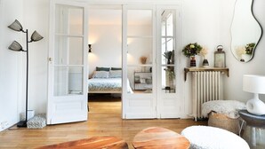 A room with a wooden floor, a bed with a white headboard, a white door, a floor lamp, a mirror, and a small table with a lamp. - Very bright. 1 bedroom Batignolles / Opera. (Paris)