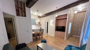 A modern living room with a sofa, a coffee table, and a TV. There is a dining area with a table and chairs, and a kitchen area with cabinets and a countertop. - Beautiful Renovated Apartment in Le Marais (Paris)