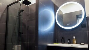 A modern bathroom with a glass-enclosed shower, a round mirror with a light, and a dark tiled wall. - New Studio at the Heart of Opera - Paris 1st (Paris)