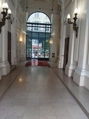 A spacious hallway with a red carpet leading to a glass door entrance, flanked by columns and wall sconces.
