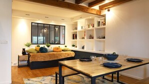 A modern living room with a wooden floor, a sofa with cushions, a dining table with plates and glasses, and a bookshelf with books and decorative items. - Spacious apartment in Batignolles (Paris)