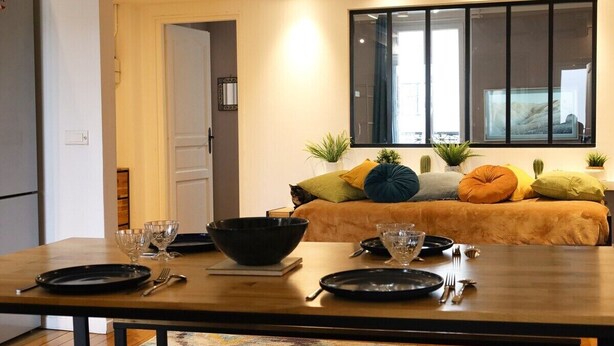 A dining area with a wooden table set for two, a sofa with cushions, and a large window with blinds. - Spacious apartment in Batignolles (Paris)