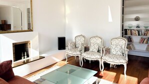 A living room with a fireplace, three upholstered chairs, a glass coffee table, and a bookshelf. - Spacious | in the Heart of the Marais | 4 people (Paris)