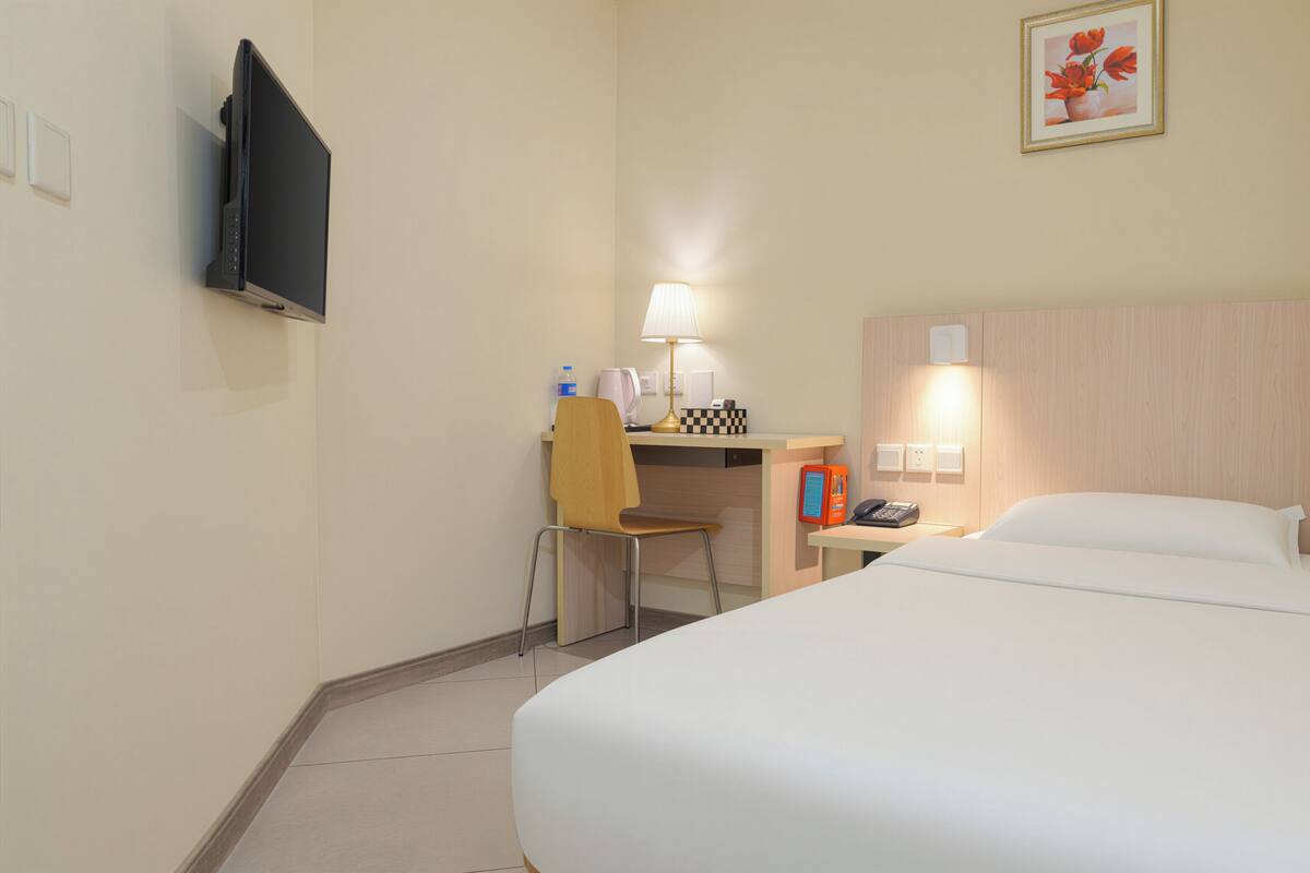 Quarto de hotel com cama, escrivaninha, cadeira, luminária e televisão fixada na parede.