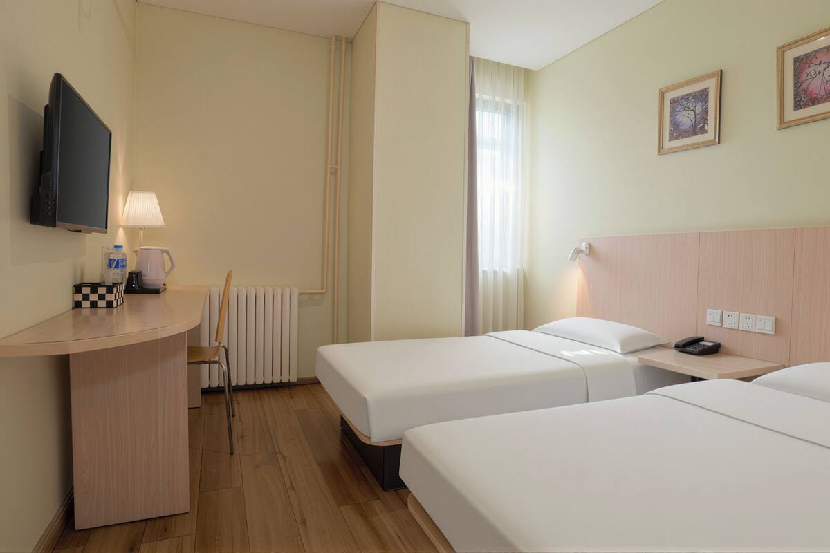 Quarto de hotel com duas camas, uma escrivaninha com abajur, televisão e radiador.