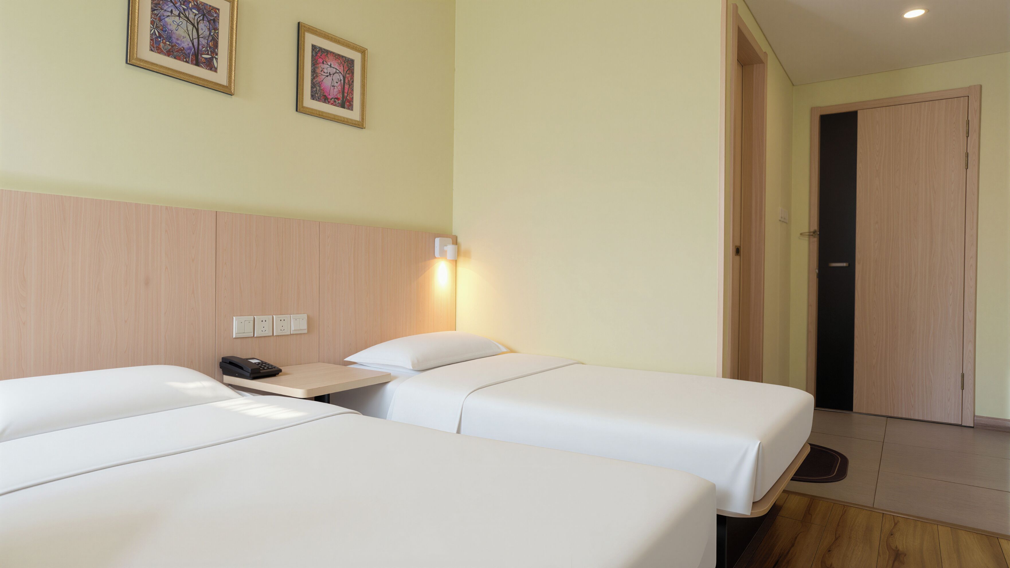A hotel room with two beds, a bedside table with a phone, a wall-mounted light, and a door.