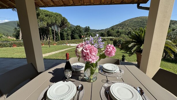 A table set for two with white plates, silverware, and a bottle of wine, set against a backdrop of a lush garden and hills.