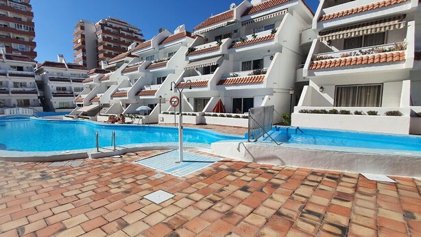 A swimming pool area with a tiled deck, surrounded by multi-story buildings with balconies and red-tiled roofs. - Las Floritas 5 - Plumeria (Playa de Las Americas)