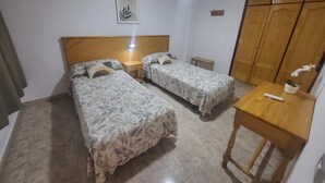 A bedroom with two beds, a wooden wardrobe, a small table, and a wall-mounted light fixture. - Las Floritas 5 - Plumeria (Playa de Las Americas)