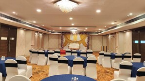 Un salón de conferencias con mesas redondas, sillas blancas adornadas con cintas azules, un escenario con fondo dorado y una araña.