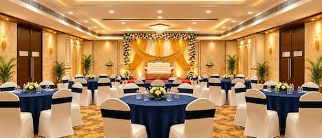 Un salón de banquetes con mesas redondas, sillas blancas y un escenario con decoraciones florales.