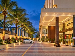 A modern hotel entrance with a paved walkway, palm trees, and a white car parked in front. - The Altruist Wagholi Hotel (Pune)