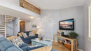 Ein Wohnzimmer mit blauem Sofa, Couchtisch, an der Wand montiertem Fernseher, einer Treppe und einer Wandmalerei mit einer Zimmerpalme.