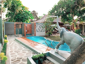 A swimming pool with a stone pathway leading to it, surrounded by greenery and a building with a tiled roof. - Kamulyan Heritage  (Depok)