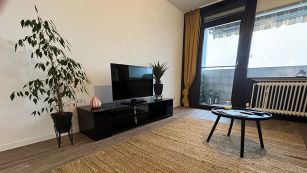 En moderne stue med et fladskærms-tv, en potteplante, et lille bord med et glas og udsigt til det fri gennem et stort vindue.