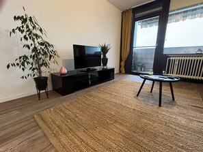 Un salon moderne avec un téléviseur à écran plat, une plante en pot, une petite table avec un verre, et une vue sur l’extérieur grâce à une grande fenêtre.